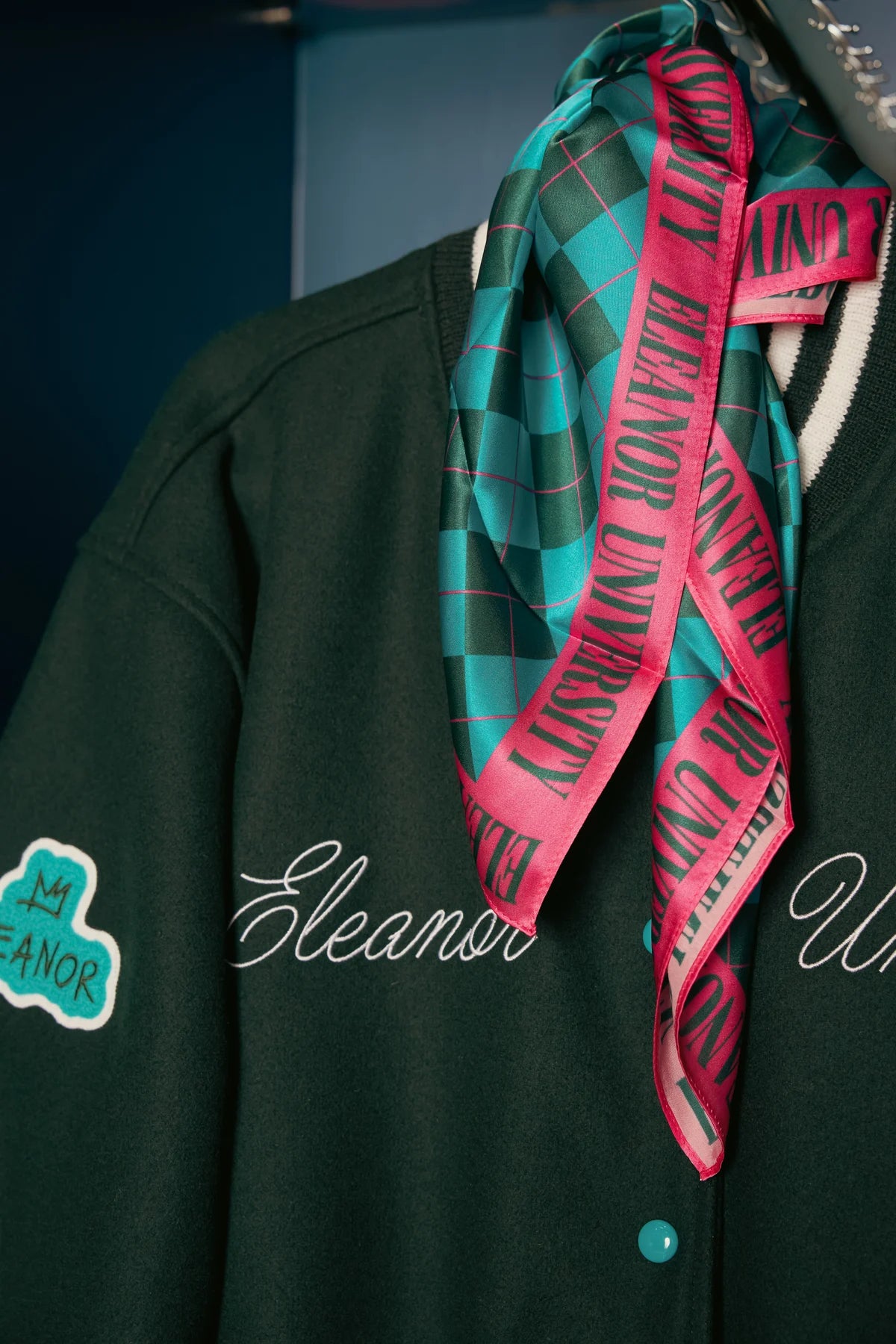 The turquoise and forest-green checked Eleanor University scarf with its pink lettered border, draped over the varsity jacket